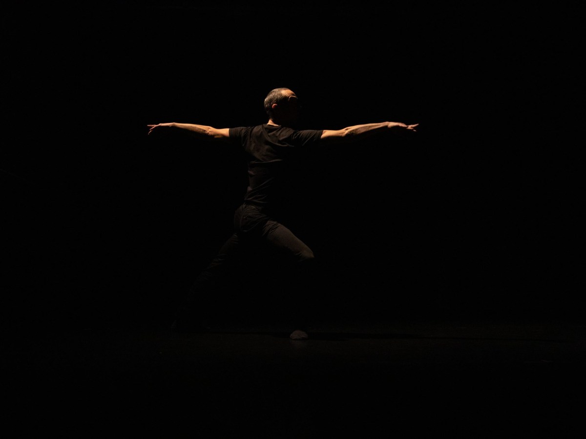 Terence Marlin, a middle-aged white man, dances in semidarkness. He is wearing dark trousers and a dark short-sleeved shirt. He is seen in profile, his arms stretched to either side.
