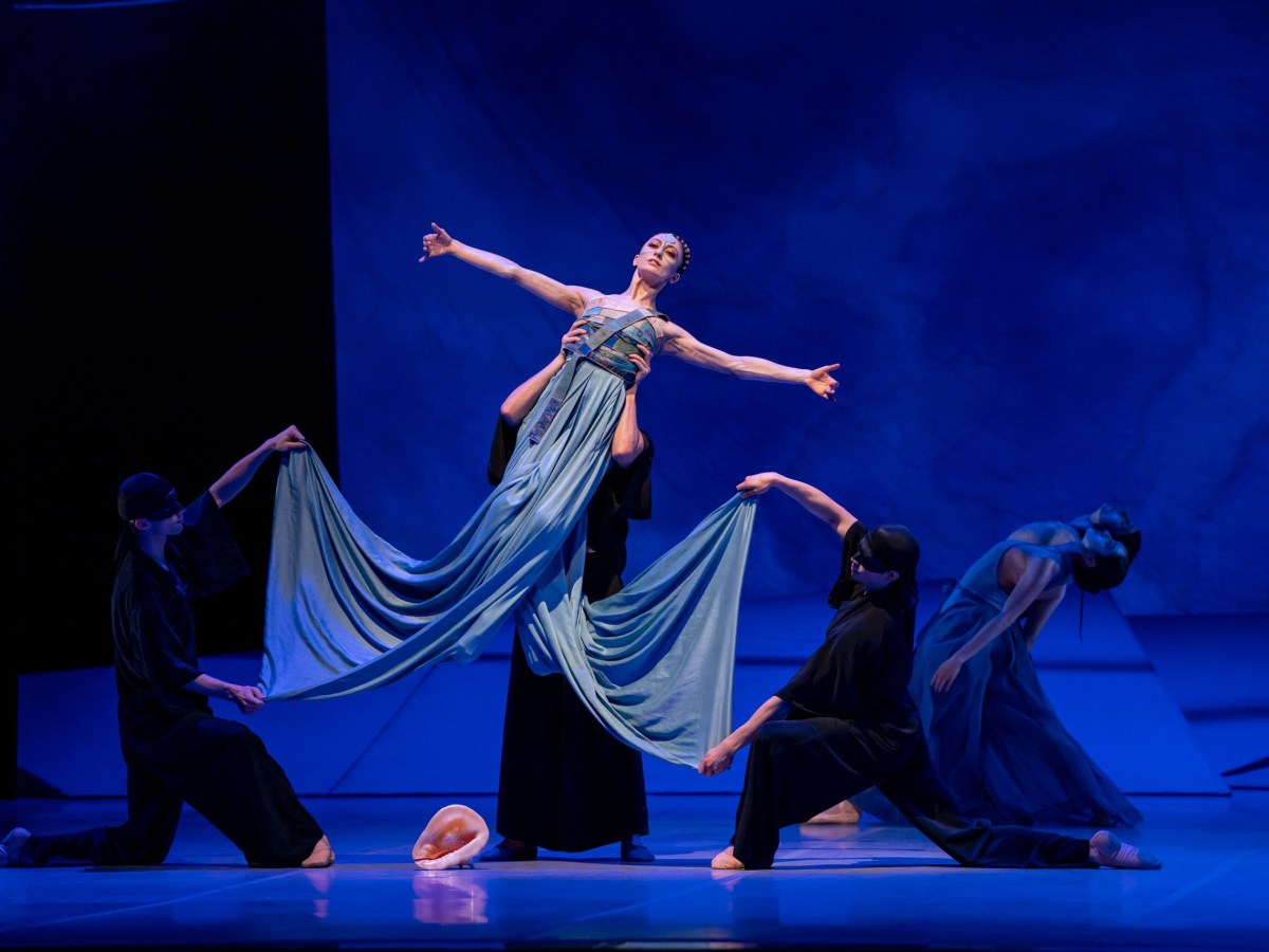 A dancer in a long light-blue bandage dress with a split skirt like long fins is held aloft by dancers in black. Her arms are outstretched to her sides.
