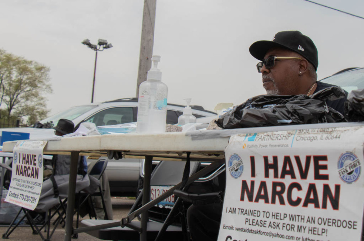 Ed Richardson wearing a baseball cap and sunglasses sits behind a folding table set up outside on Pulaski Road in Chicago. A sign taped to the table reads I Have Narcan and provides information about the outreach programs that do public programs for opioid users
