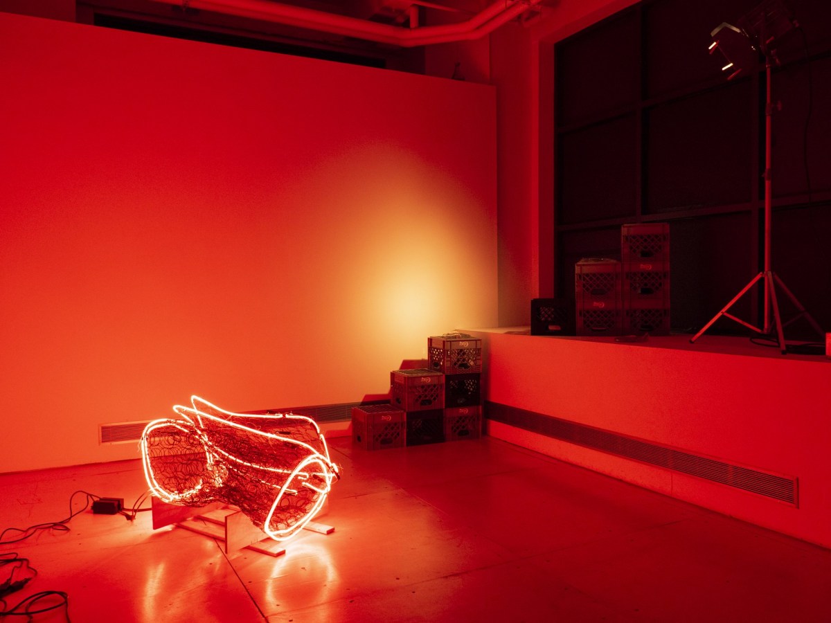 The room is tinted by a bright red neon light. The light wraps around a gnarled wire bed frame, in a bundle on the floor. A stack of milk crates sits off to the side, and a bright stage light shines down on the whole scene.