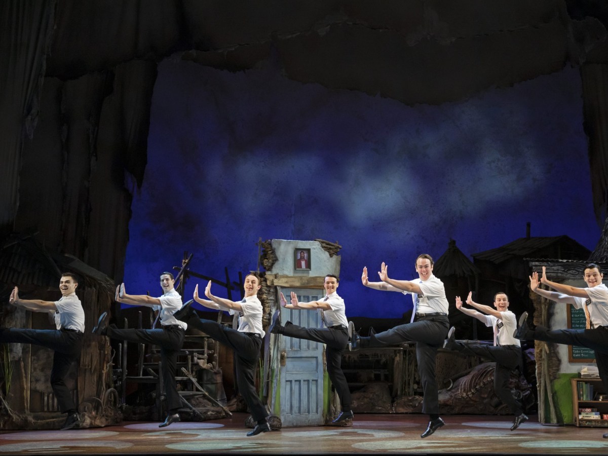 Seven cast members of The Book of Mormon dance in a row, facing in profile to the left of the photo. Their arms are stretched in front of them, and each of them has one leg kicked out straight in front. They are wearing Mormon missionary uniforms (black trousers, short-sleeved white shirts, ties, black shoes).