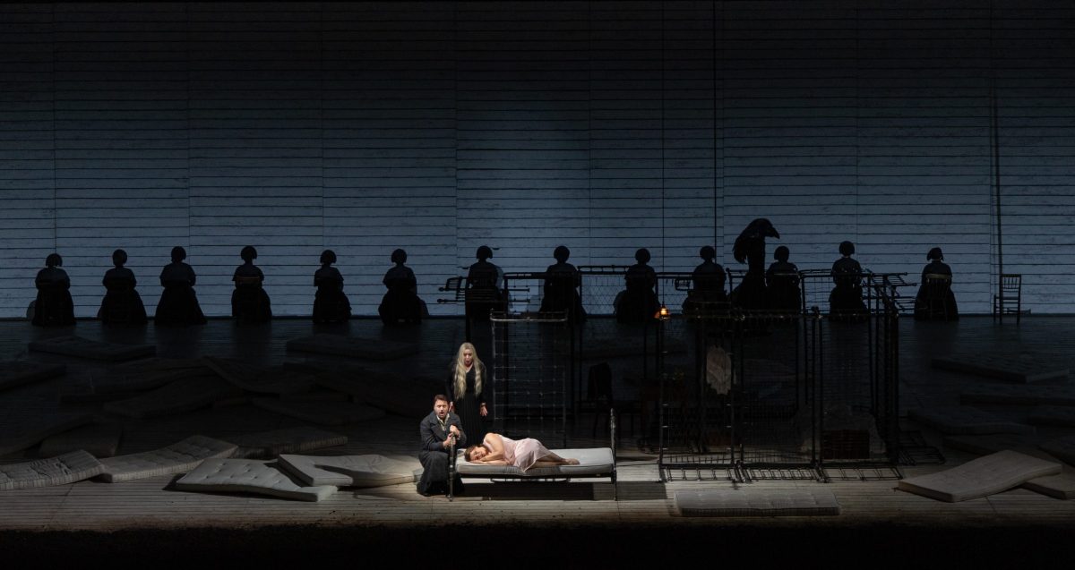 A line of figures silhouetted in the background with their backs to us. A woman in a light-colored slip lies on an iron frame bed in the center front. A man kneels next to her at the head of the bed on the left, and another woman in a black dress stands behind her. There is a row of cages on the right and more mattresses strewn over the floor.