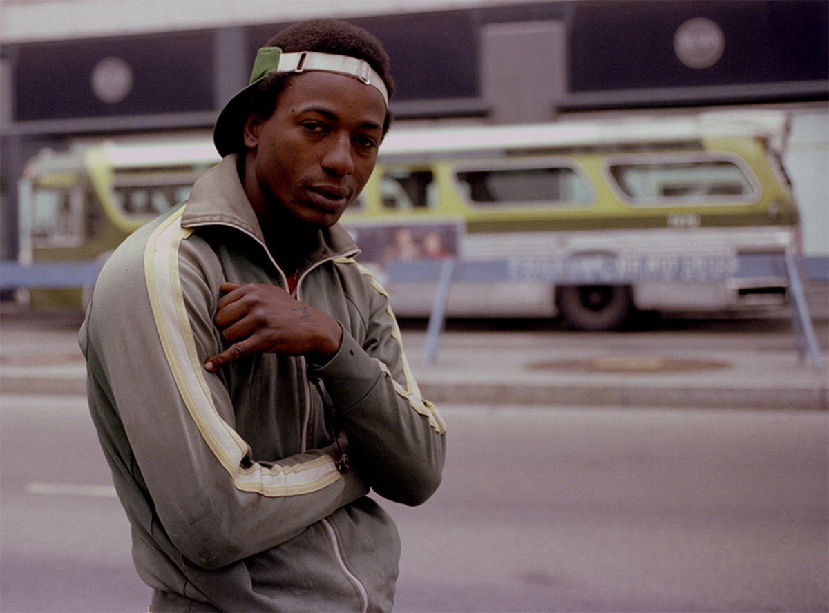 a Black man wearing a backward visor on his head and a warm up jacket with a stripe on the arm; a green cta bus is visible in the background