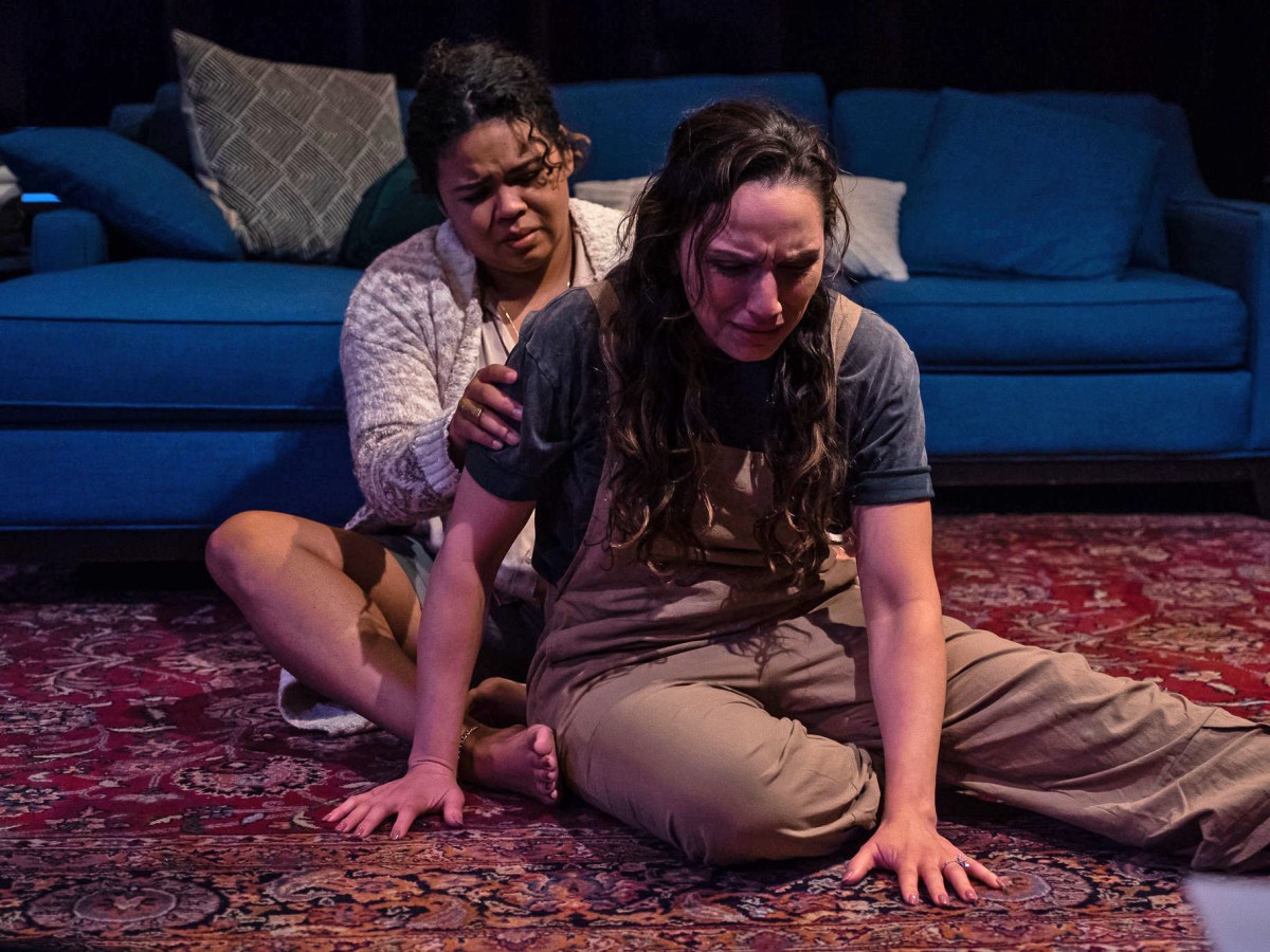 Two Latinx women sit on the rug in a living room. A blue couch is behind them. Marc (Melissa DuPrey) sits behind Cynthia (Lisandra Tena), who seems to be in emotional distress. Marc has her hands on Cynthia's back.