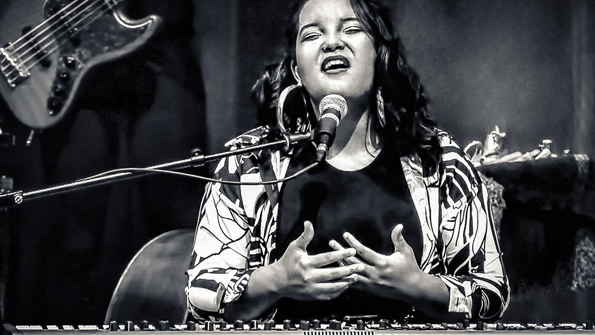 a black-and-white photo of Alexis Lombre, facing the camera with her eyes closed as she sings into a microphone while seated behind a keyboard, her fingers interlaced and hands palm-up above the keys