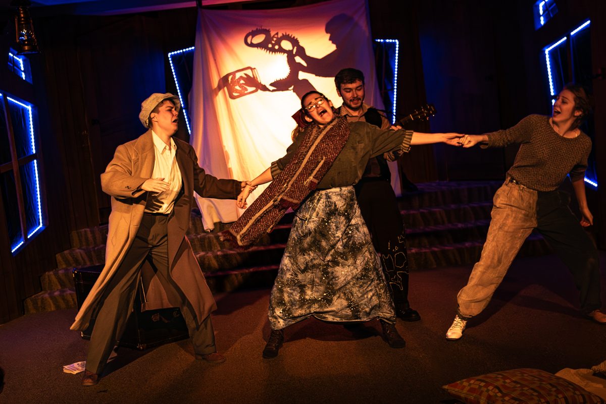 Four people are onstage. A person in the middle is being pulled by the hands in opposite directions by two other people. Behind them is a fourth person, and behind all of them is a screen with a shadow puppet of a scary-looking creature.