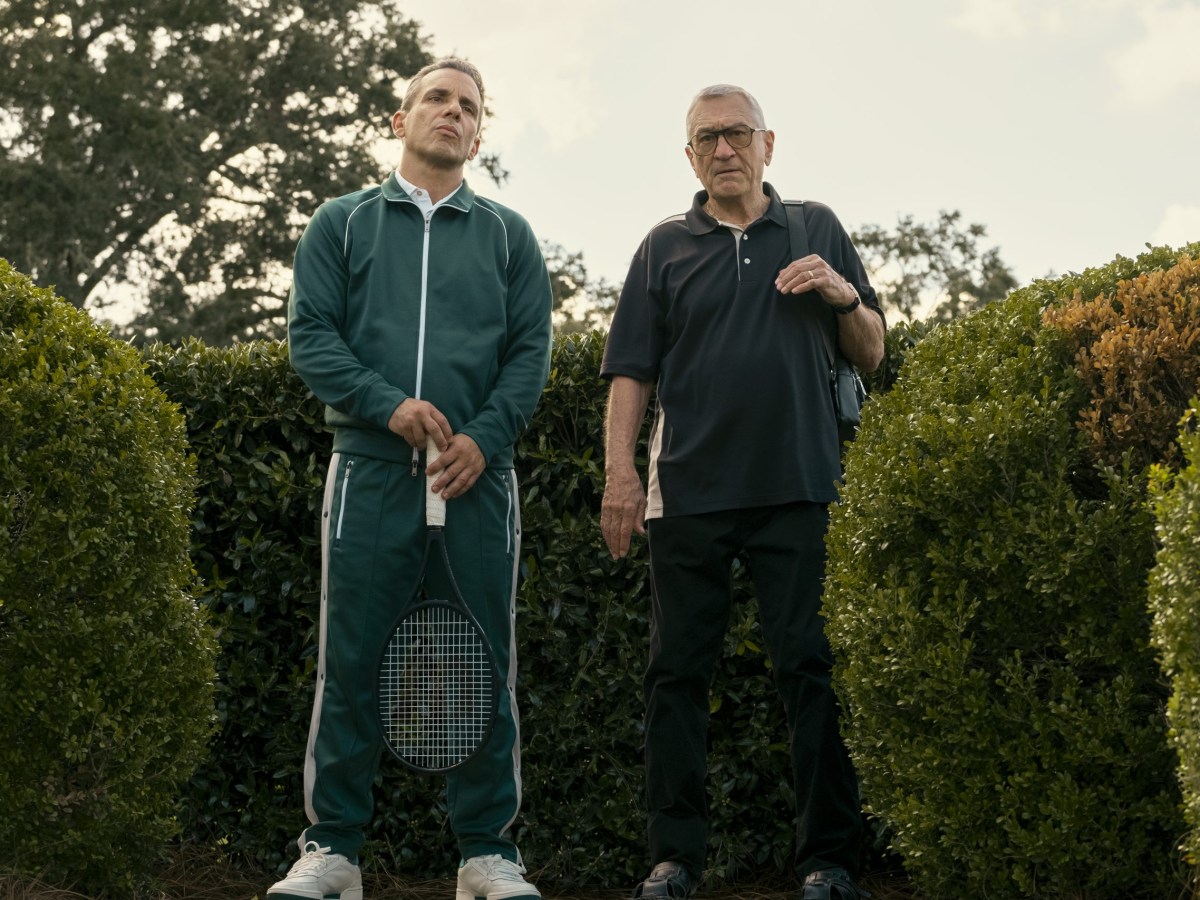 two men stand in front of hedges, one holding a tennis racket