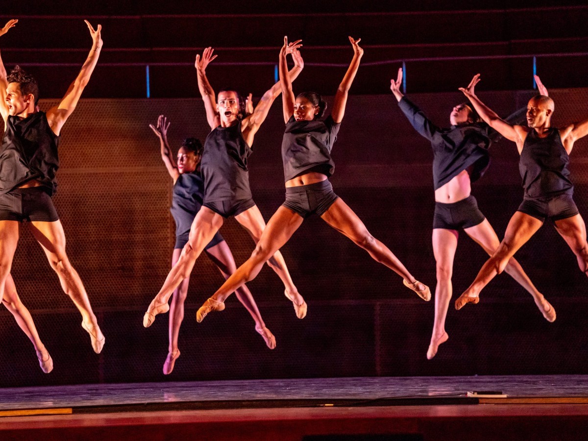 A group of dancers in black shorts and tank tops are shown leaping in the air with their arms stretched above their heads.