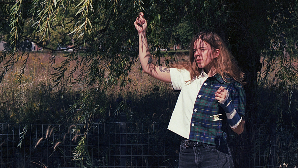 Ash Dye stands near a wire fence on a sunny day in what looks like parkland, her right arm up in the leaves of a low-hanging tree behind her and her left hand (in a wrist brace) held near her chest.