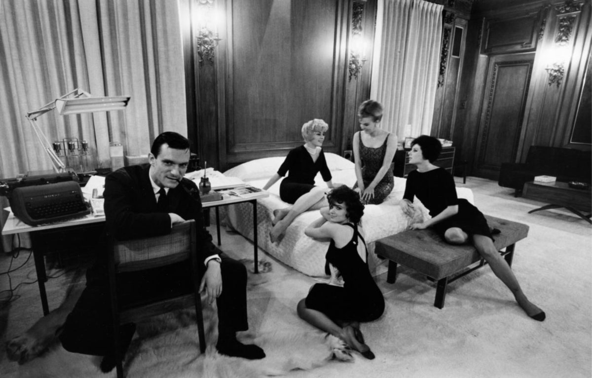 A black-and-white image by Art Shay of Hugh Hefner in his bedroom. Hefner is wearing a dark suit, seated at a desk with a typewriter, and four young women pose on the bed.