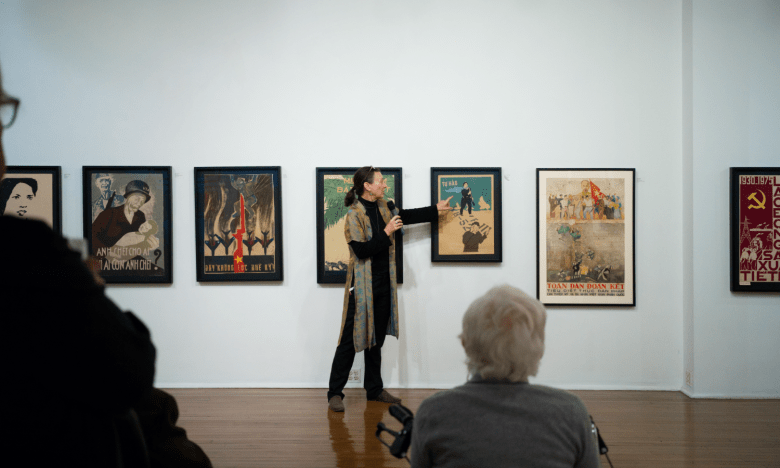 A woman with a microphone stands in front of a gallery wall hung with Vietnamese posters. She is pointing to one of the posters, an audience can be seen in the foreground listening to her.