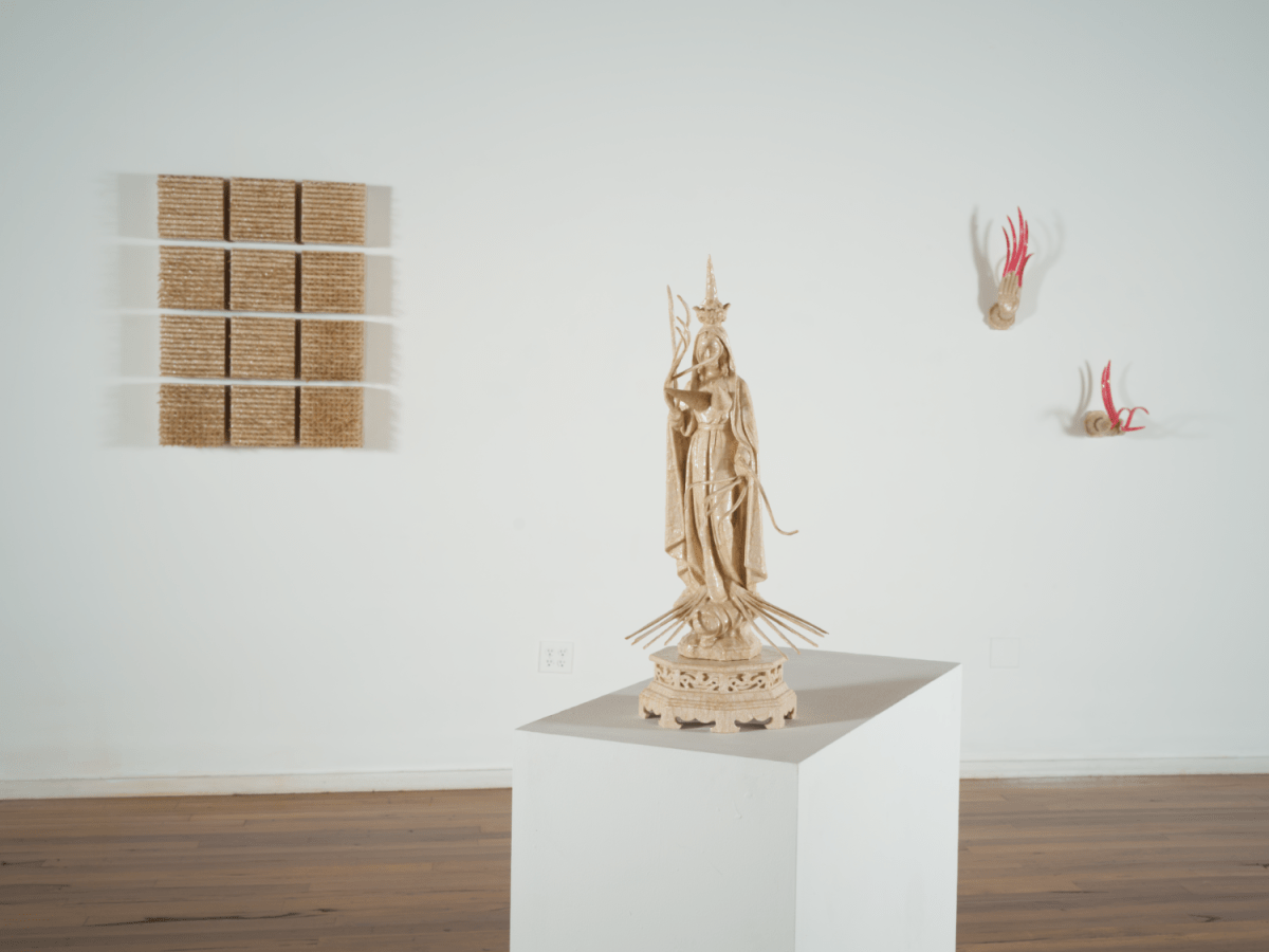 On the wall in the background, a grid of 12 boxes of sharp wooden nails protrude from the wall. Near it are two feminine hands extending from the wall, with long, threatening pink fingernails. In the foreground is a beige sculpture on a plinth of a deity with long fingernails.