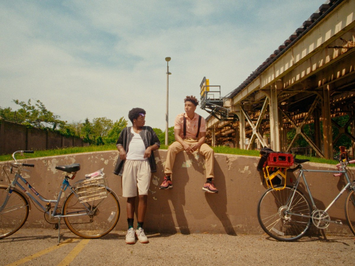 two young Black people sit or lean against a low concrete wall with their bikes under elevated train tracks