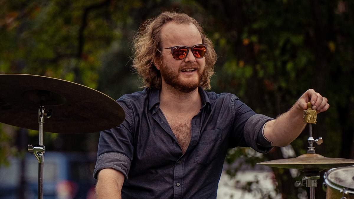 a trap-set drummer wearing sunglasses plays on a lawn, and he's reaching out toward what appears to be a small bell on top of his hi-hat stand