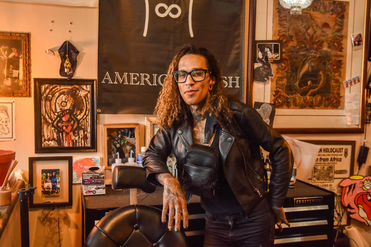a tattooed Black woman with black glasses and a leather jacket poses inside a tattoo studio