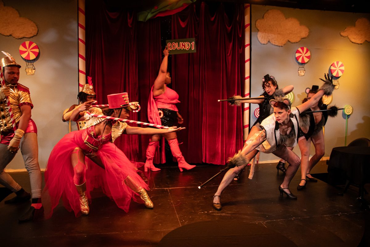Three performers dressed as burlesque versions of toy soldiers stand left, facing off against three performers dressed as burlesque mice. A person in the background between them holds up a placard reading ROUND 1.