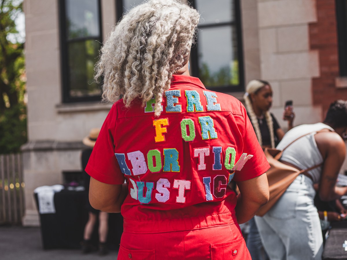 A woman stands with her back to the camera wearing a jacket that reads "here for abortion justice"