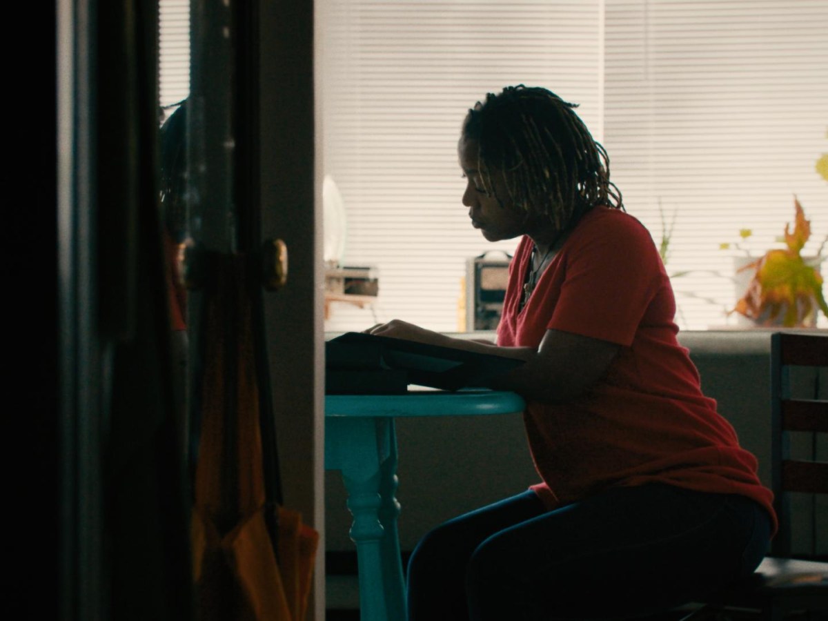 A Black woman sits at a blue table, appearing to look at documents. She is seen through an open door in low light.