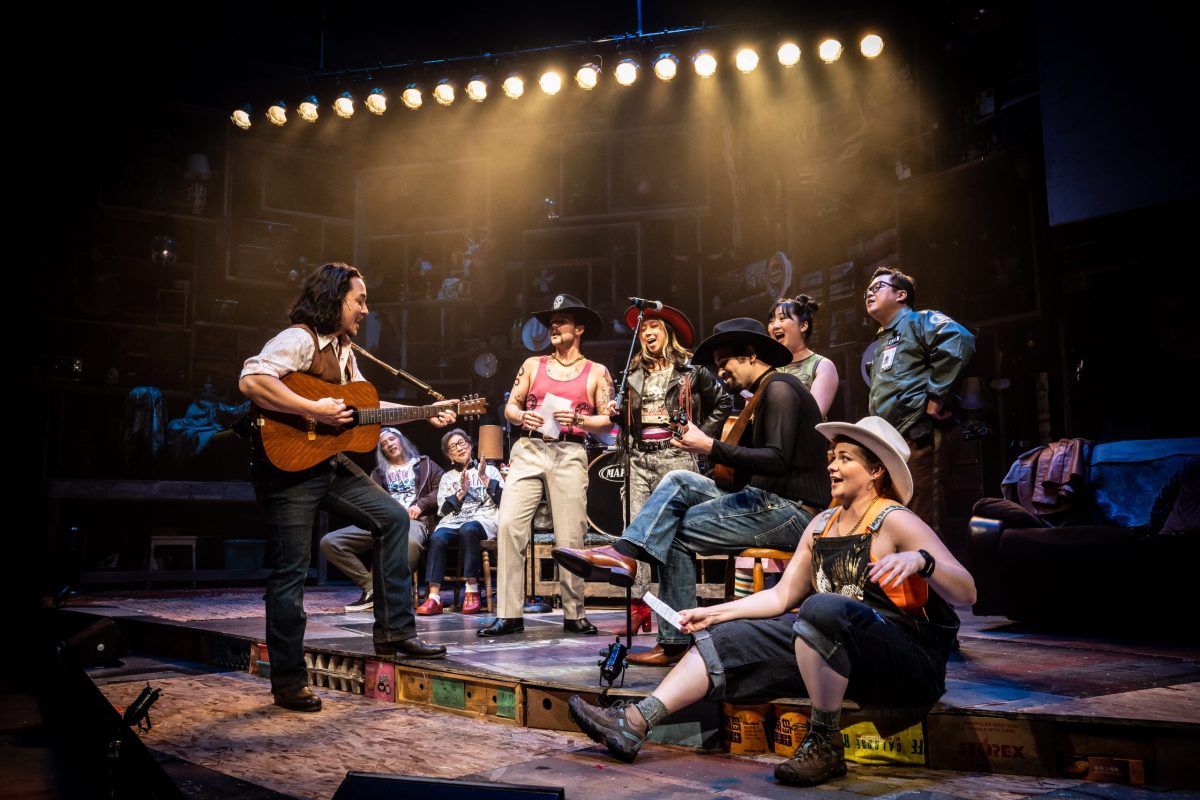 The cast of Lucy and Charlie's Honeymoon at Lookingglass Theatre stand and sit center stage, with several of them playing musical instruments.