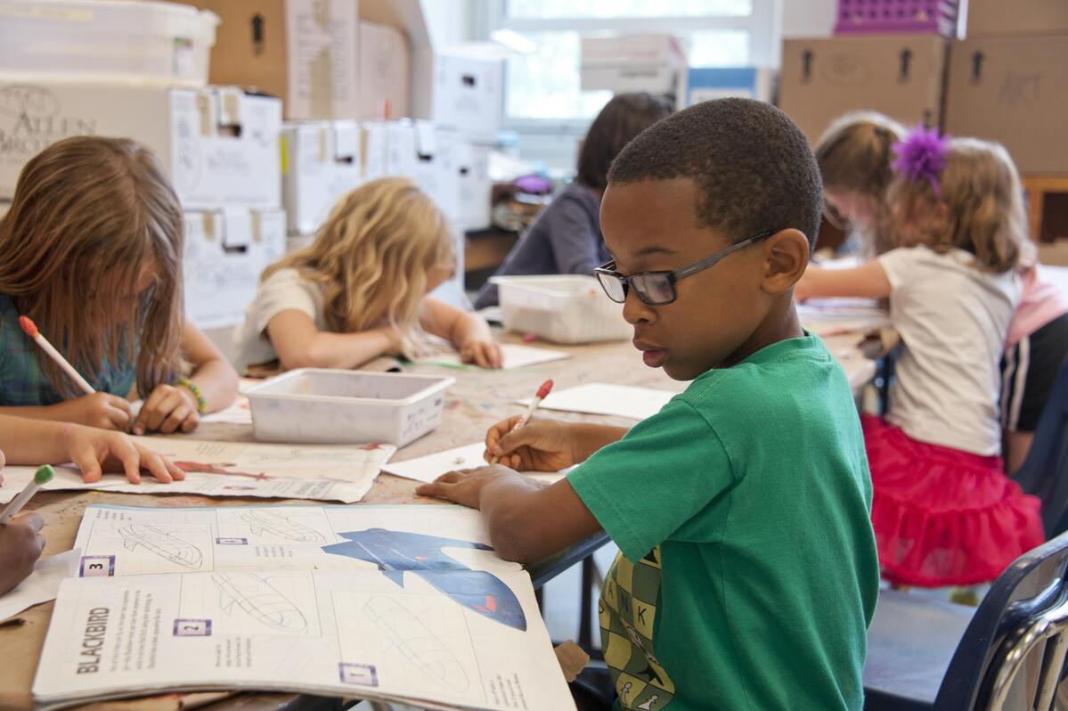 Elementary school aged students in a classroom, a Black boy in a green tshirt, wearing glasses, is in the front of the photo and is doing a workbook page.