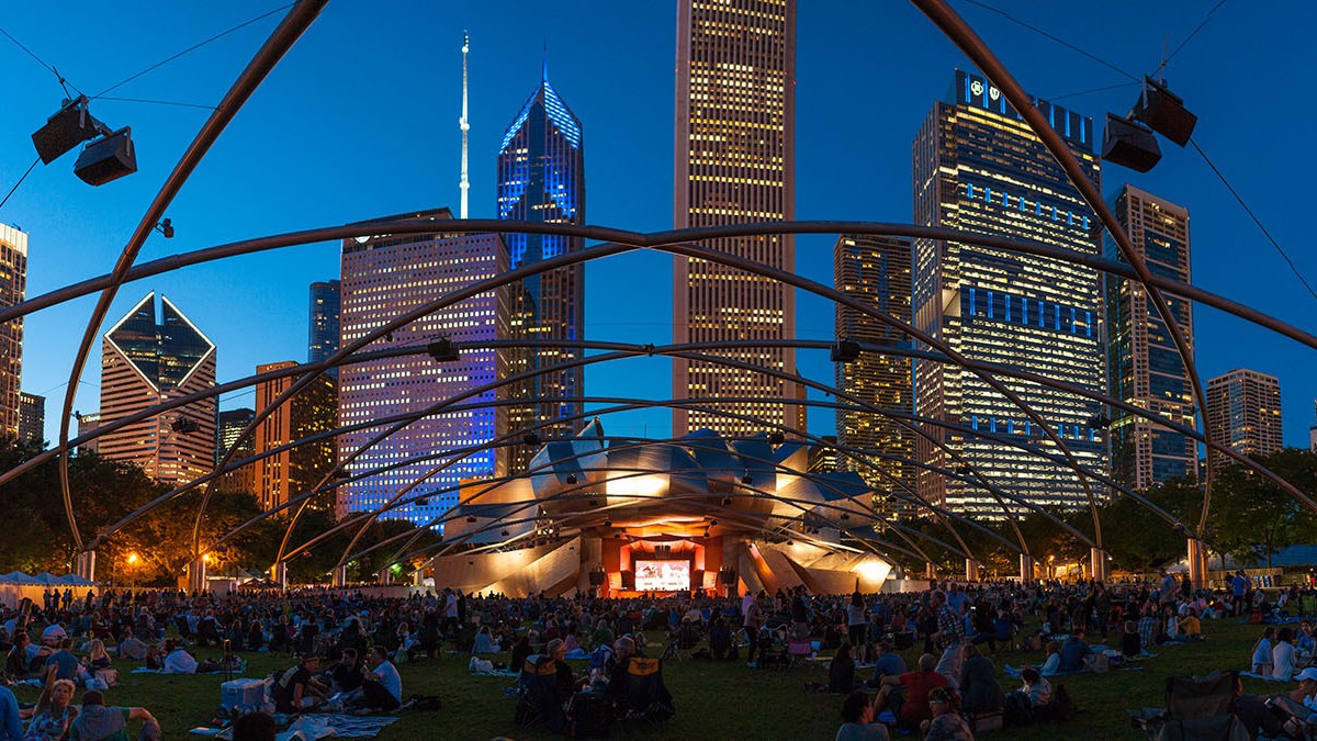 a distant view at dusk of the brightly lit stage of Pritzker Pavilion and the lattice of overhead speakers in the park, taken during a previous Jazz Festival show