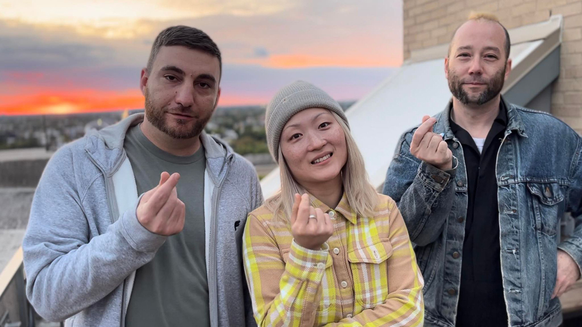 The three members of Chicken Happen face the camera on a rooftop with the sunset over the city in the background, and they're all making a hand gesture that looks like they're holding something between their thumbs and index fingers