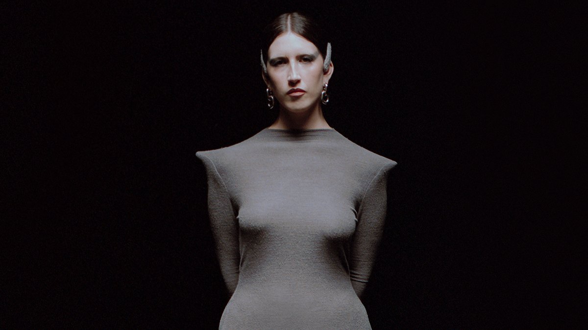 Claudia Ferme, aka Claude, stares into the camera in a flat angle against a blank black background wearing a close-fitting sci-fi-style gray dress with pointed shoulders