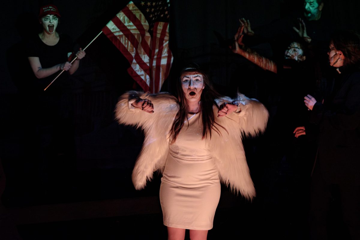 A woman in white dress and fur coat, wearing white makeup, stands center in dim light. Behind her we see figures in black clothing and white makeup. The one on the left is wearing a red MAGA hat and waving an American flag.