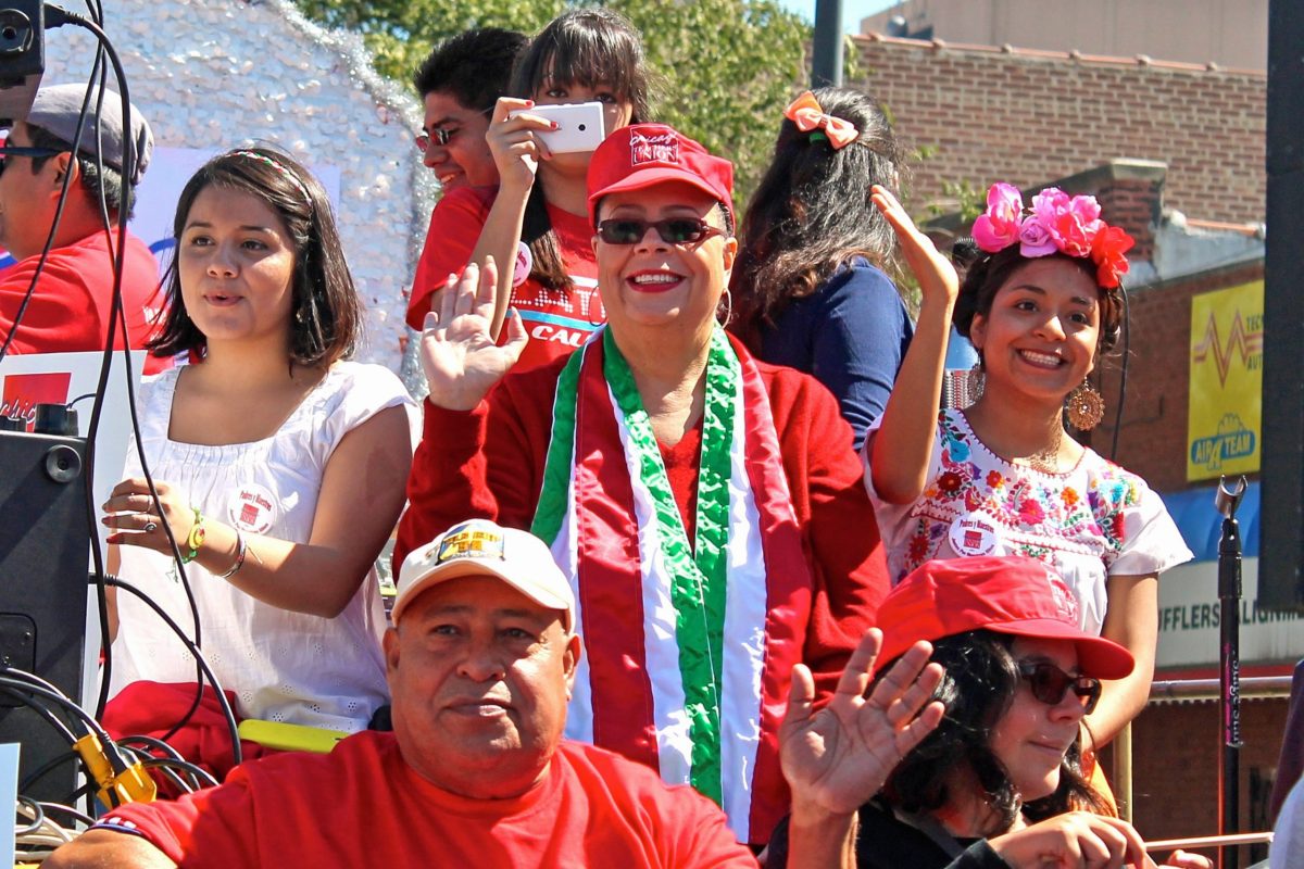 Karen Lewis wearing a red cap and red shirt standing in the middle of several parade participants on the Chicago Teachers Union float in 2014's Mexican Independence Day Parade in Chicago