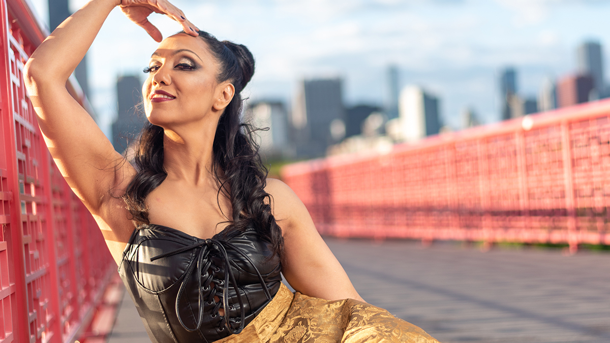 a seated Czarina Mirani leans against a red-lacquered latticework bridge railing in Chinatown's Ping Tom park in a black and gold gown, with blue sky and the Chicago skyline visible behind her