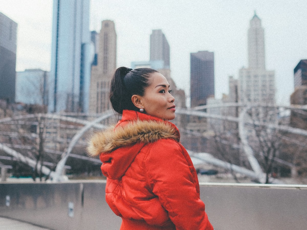 A Black woman in a red winter parka looks over her right shoulder and past the camera while standing in front of sculptures at Chicago's Millennium Park
