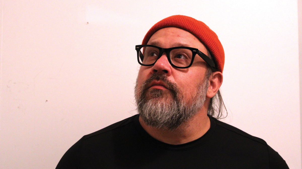 a head-and-shoulders photo of a bearded man in a red cap and dark-framed glasses, looking up and away