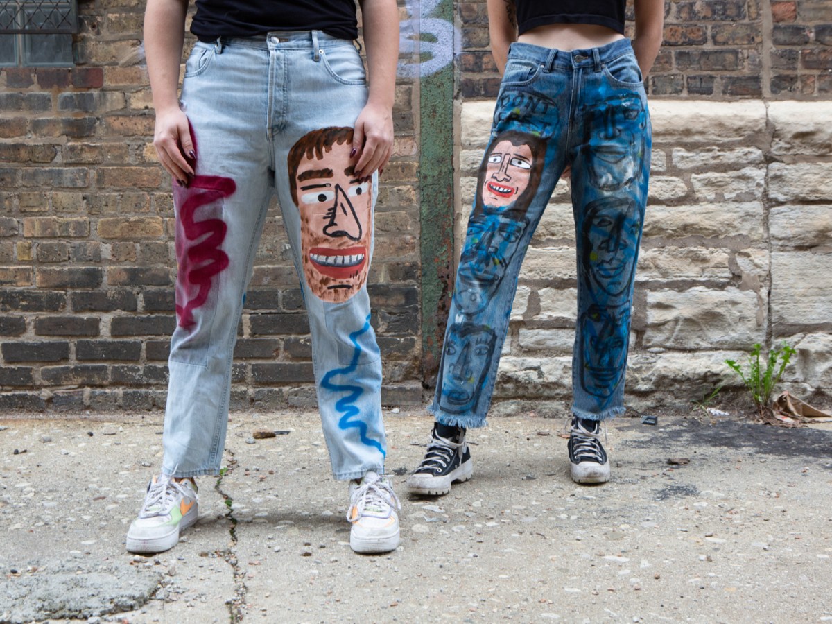 Two models are seen from the waste down. They are standing in an alley with a building behind them. Both wear jeans painted with faces and other designs.