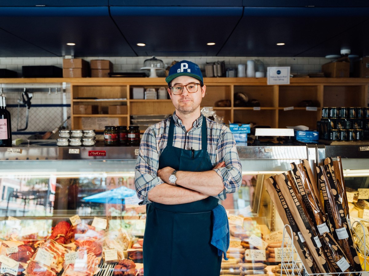 an aproned white man stands with his arms crossed in front of a butcher case