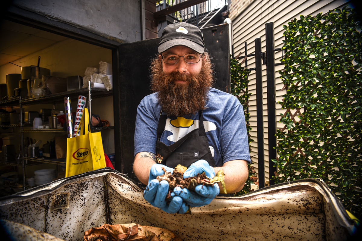 a white man with a scruffy brown beard, glasses, hat, and apron holds up a handful of compost