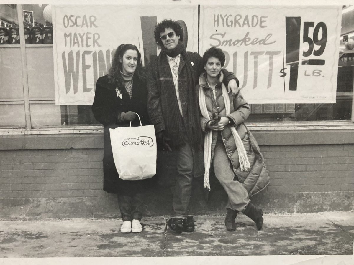 Three people stand in front of a grocery store window in this black-and-white photo. Two women flank a man. The woman on the left is holding a bag with the legend Econo-Art emblazoned on it.