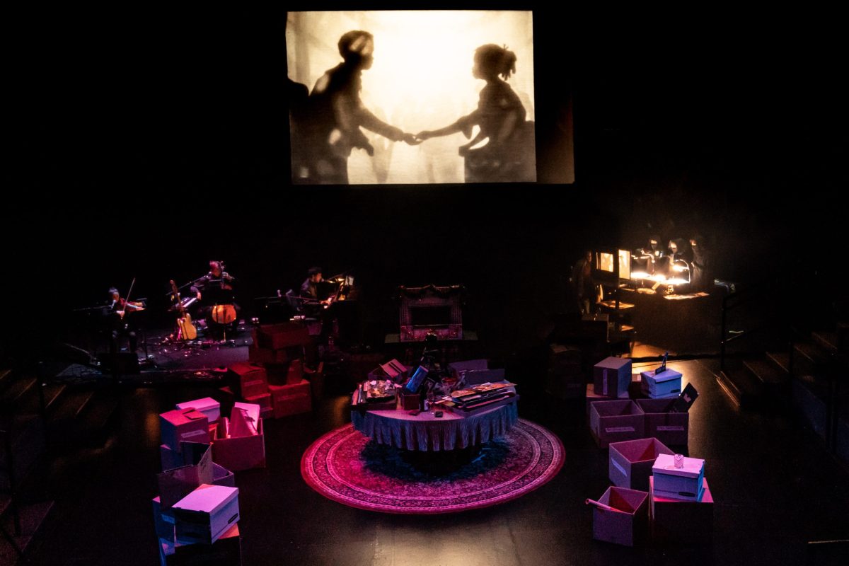 In the background we see two shadow puppets in profile, reaching out their hands to each other. In the front we see a stage with a dining room table, packing boxes, and a group of musicians in the upper left.