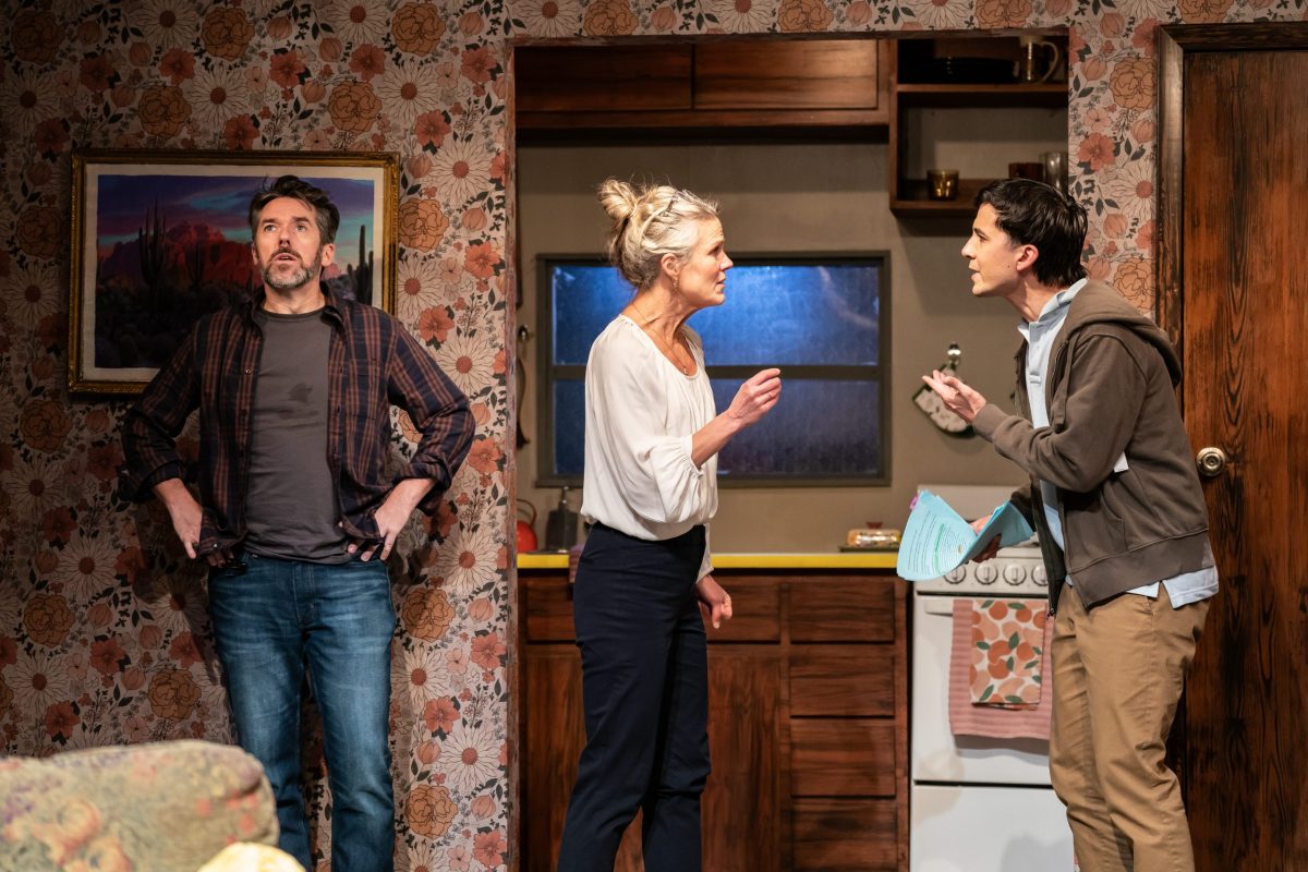 A man with a beard stands left, his hands on his hips. Next to him is a woman who is facing off against a younger man, who stands right. Behind them is a wall covered with flowered wallpaper and a glimpse of a 1970s-style kitchen.