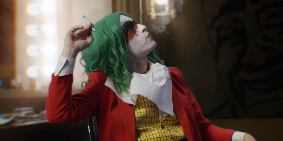 a Joker figure with white face paint, green hair, and a red and yellow outfit sits and smokes a cigarette