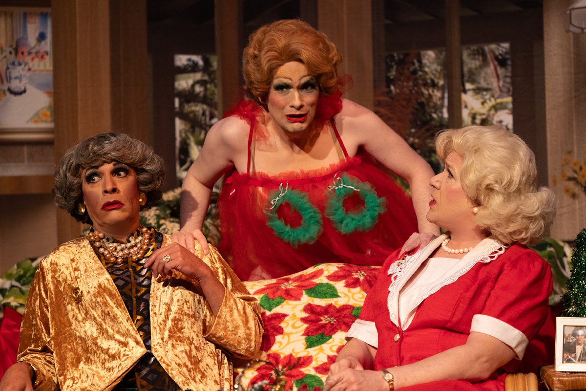 Three performers dressed as Dorothy, Blanche, and Rose from The Golden Girls. Dorothy is seated on a couch on the left, Rose is on the right, and Blanche stands between them in the back, with two small Christmas wreathes encircling her breasts.