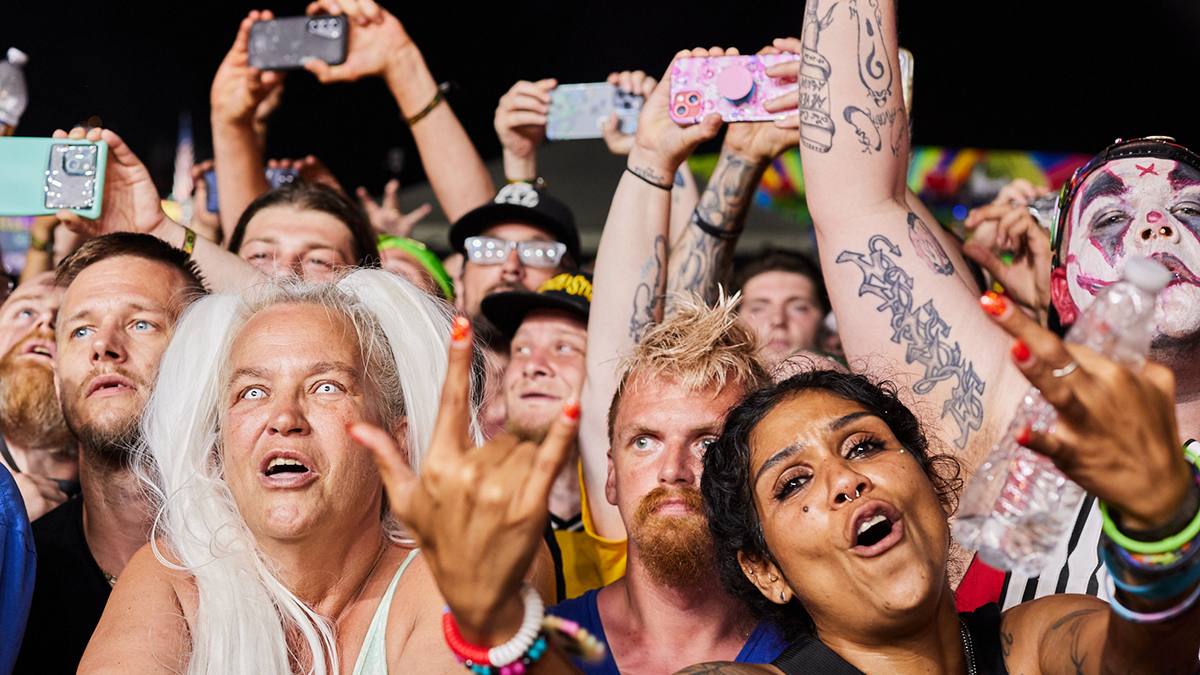 a view into a festival crowd from right in front of the stage, with lots of people holding phones in the air; you can see a white-haired woman with light-colored contact lenses, someone wearing white, black, and red juggalo-style clown face paint, and lots of tattoos