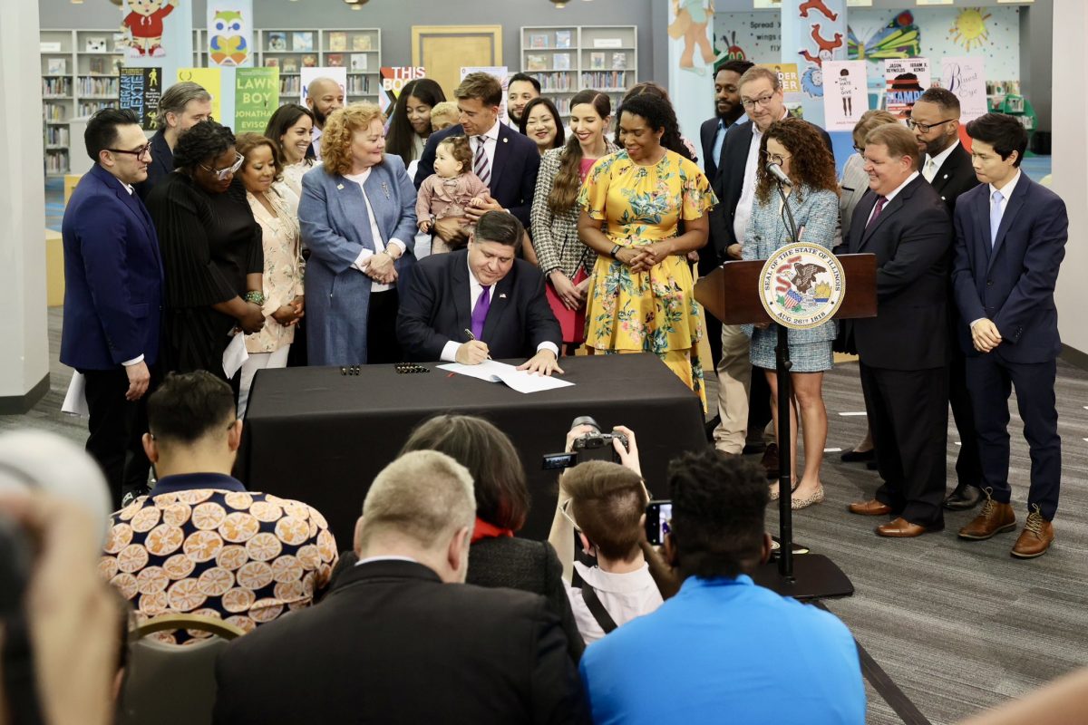 Governor J.B. Pritzker sits at a library table signing a bill, surrounded by supporters.