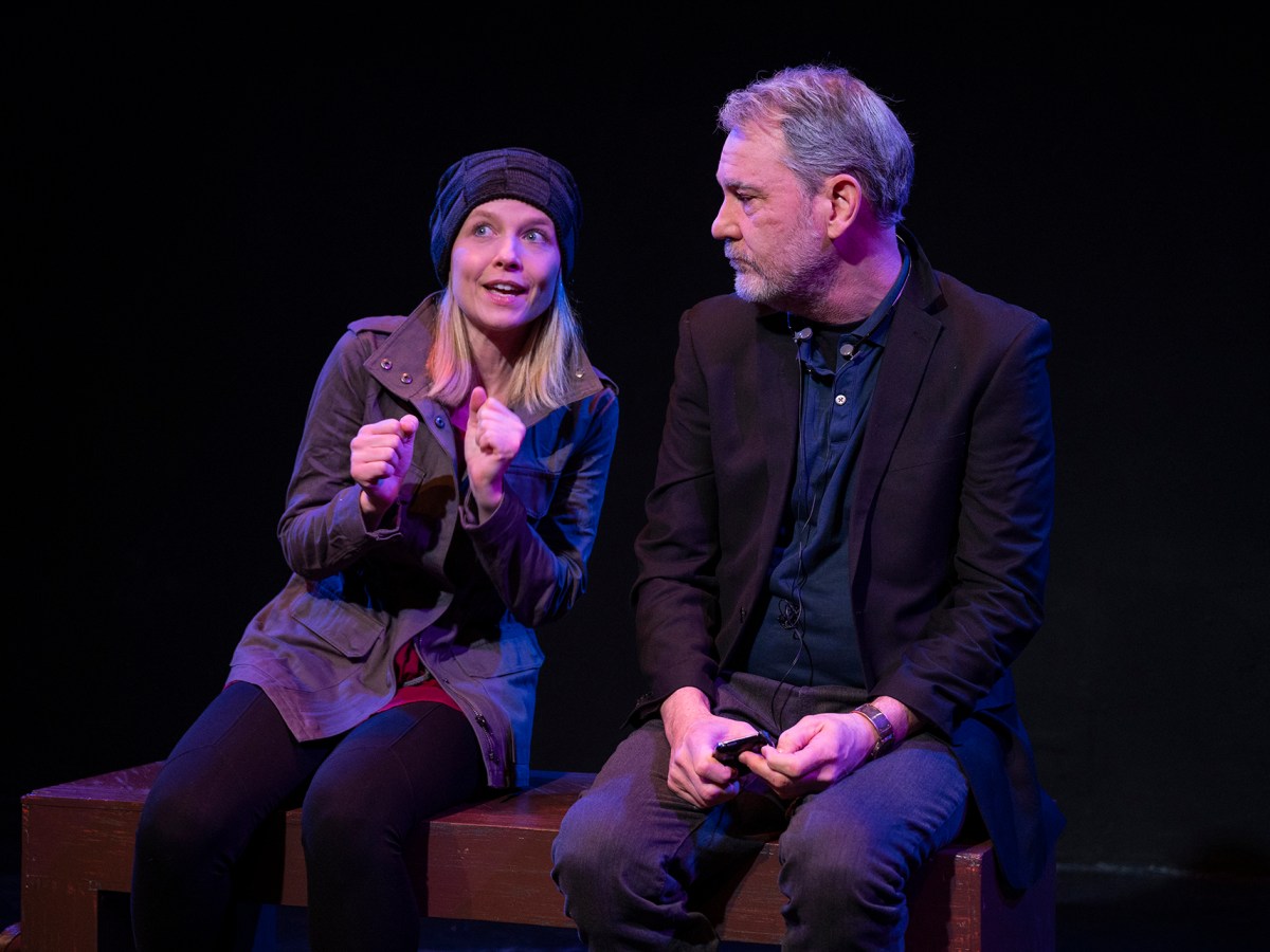 A blonde woman sits left, wearing a knit cap and a light outer coat. Her hands are balled up in excitement and she smiles as she talks. An older man with gray hair sits looking at her, his hands in his lap and holding a cell phone. He is bearded and wears dark trousers, jacket, and a blue shirt.