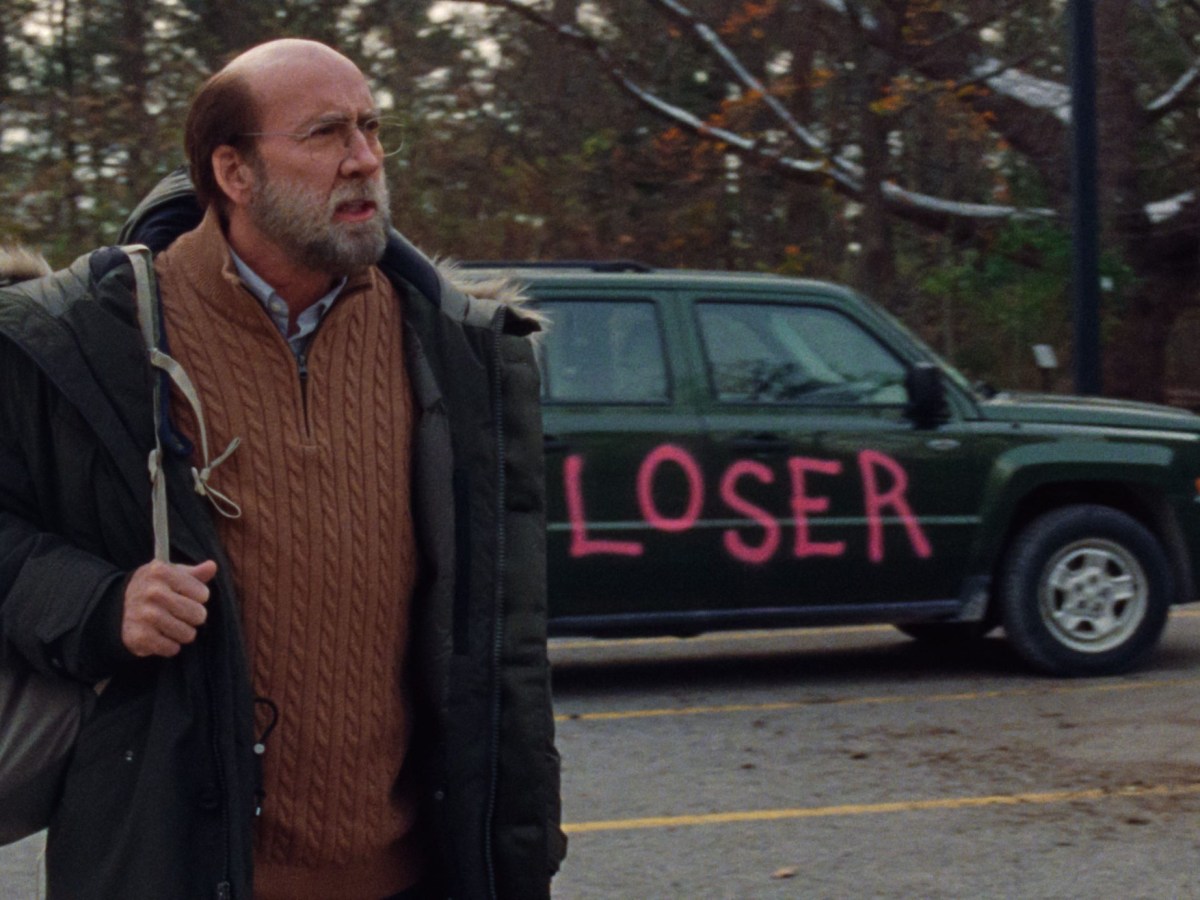 a bald white Nicolas Cage stands near a car with the word Loser spray painted on it