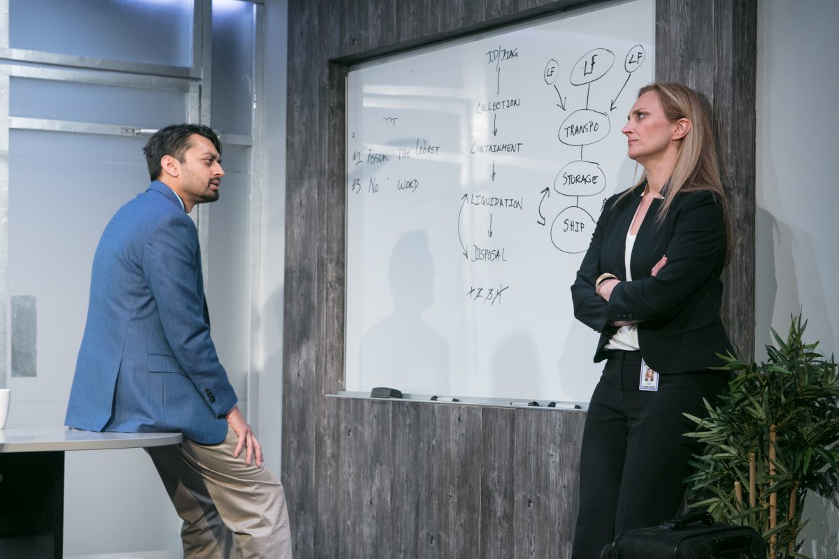 A Brown man in a blue jacket stands leaning against a desk on the left. A white woman in a black suit stands right. A whiteboard on which words are scribbled is on the wall between them
