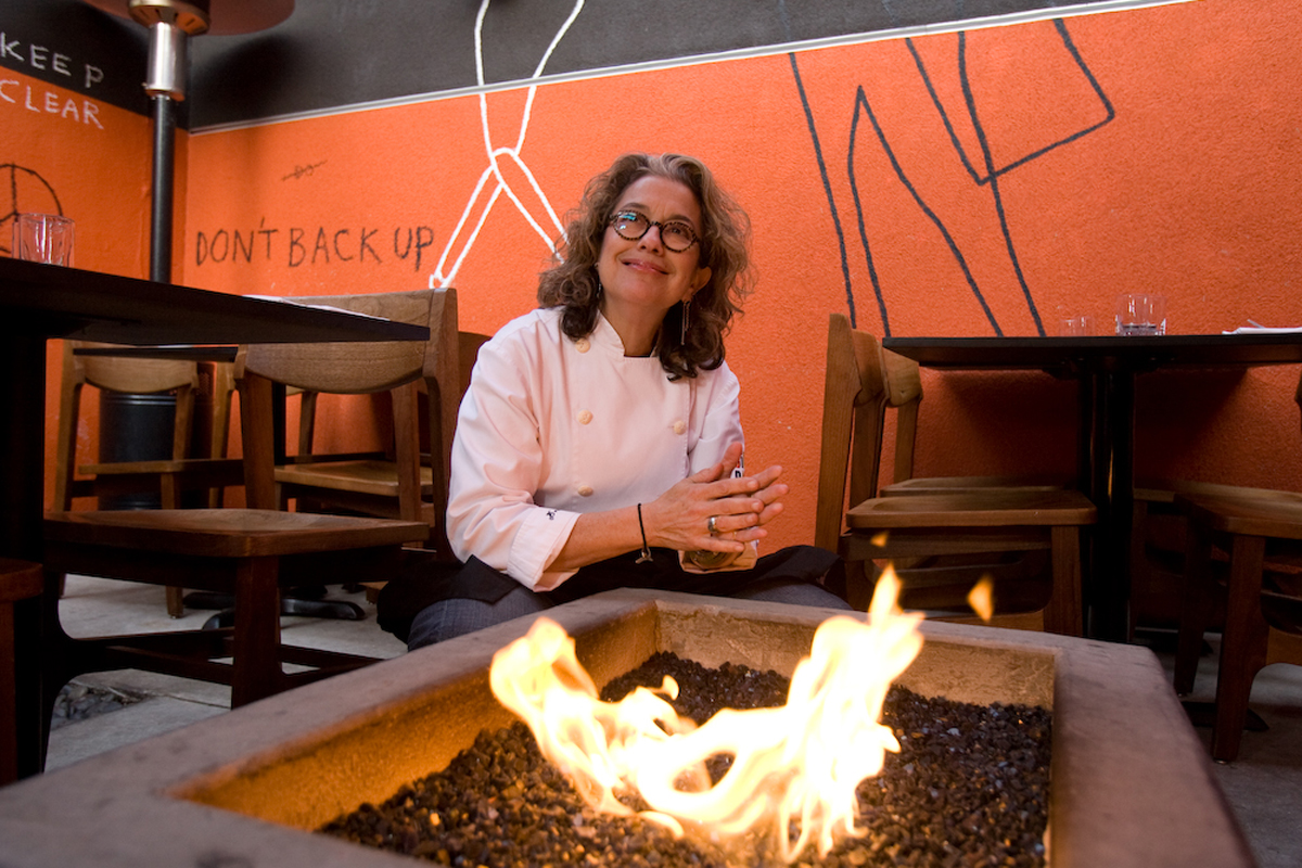 a female chef kneels by a small fire pit in an orange-walled restaurant