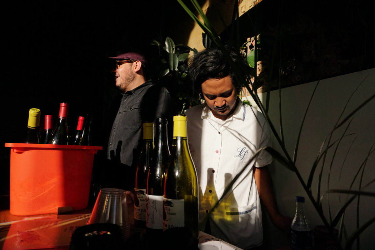 two men stand at a counter with wine bottles, dark shadows and bright sun contrasting sections of the photo