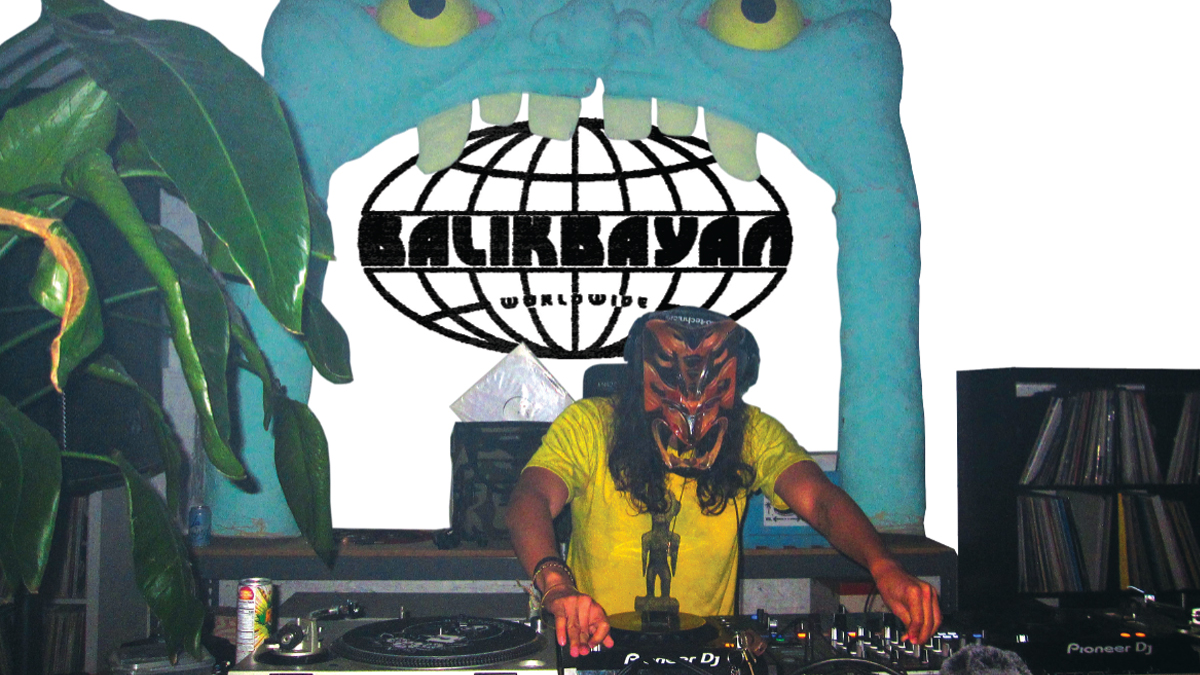 a man in a yellow T-shirt and scowling wooden-looking mask DJs on two turntables on front of a large blue monster mouth into which the Balikbayan Worldwide logo has been matted