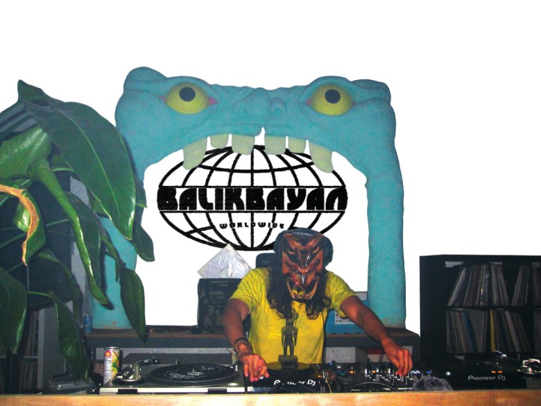 a man in a yellow T-shirt and scowling wooden-looking mask DJs on two turntables on front of a large blue monster mouth into which the Balikbayan Worldwide logo has been matted