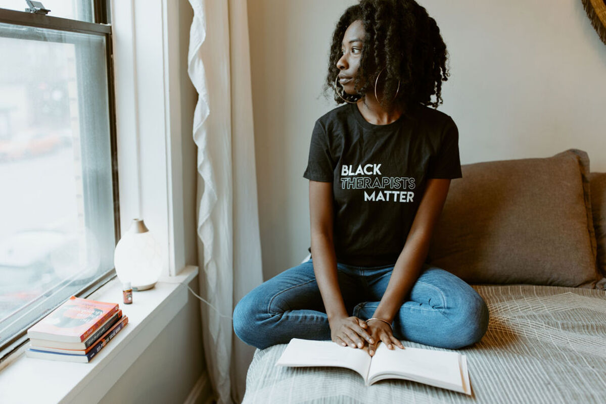 Ammie Kae Brooks sits on a bed looking out a window. Brooks is wearing jeans and a black t-shirt that reads Black Therapists Matter